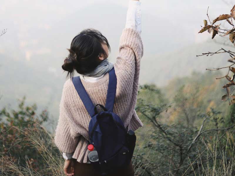 Young woman with a backpack facing a landscape and reaching up